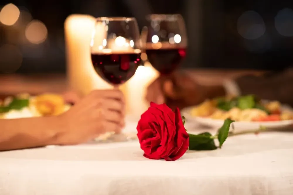Hands toasting red wine over a rose at a romantic dinner.