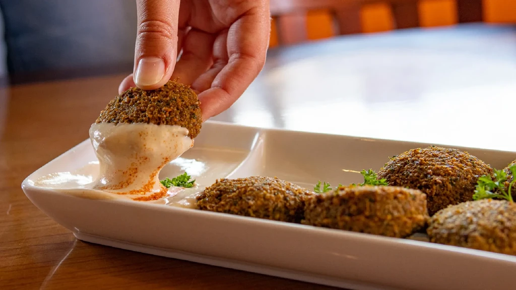 Hand dipping crispy falafel ball into white sauce on a platter.