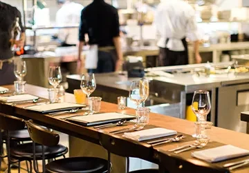 Set wooden dining counter, blurred open kitchen with working staff.