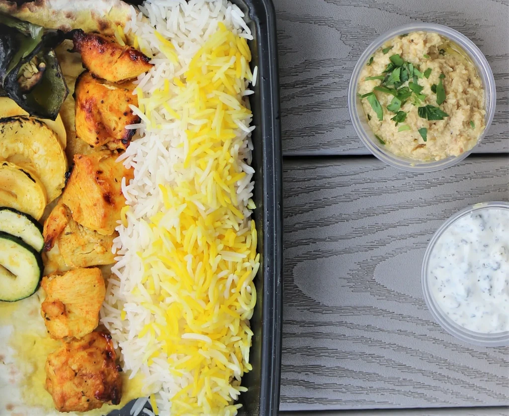 Grilled chicken, rice, vegetables, and dips in a black take-out container.