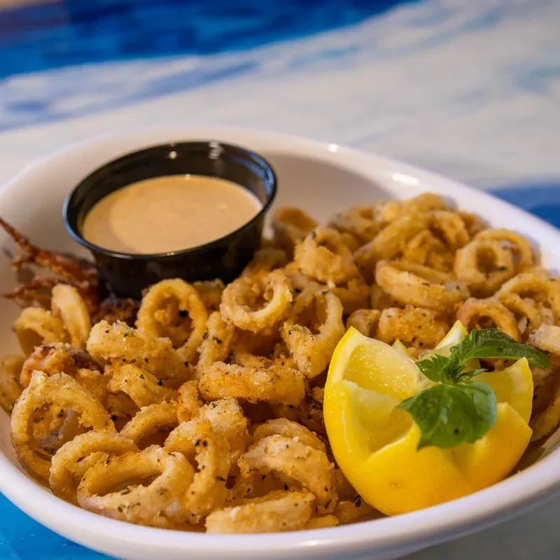 Golden fried calamari, dipping sauce, lemon, and basil in a white bowl.