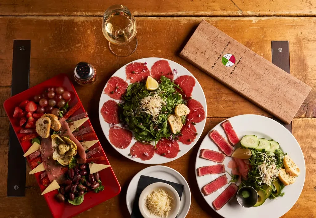 Rustic wooden table with Italian charcuterie, carpaccio, seared tuna, and wine.