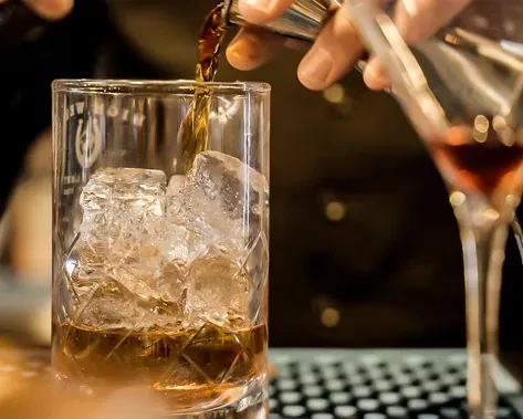 Bartender's hand pours amber liquid into an ice-filled glass.