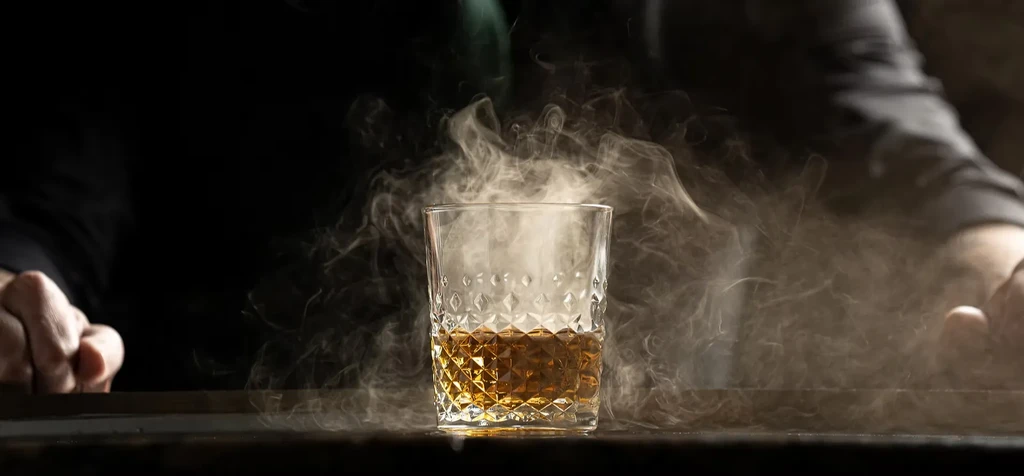 Ornate glass with amber liquid, surrounded by smoke, and visible hands.