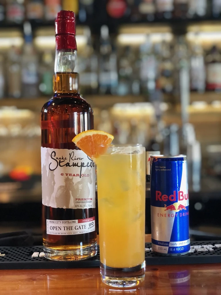 Cocktail, whiskey bottle, Red Bull can on a dark wooden bar.