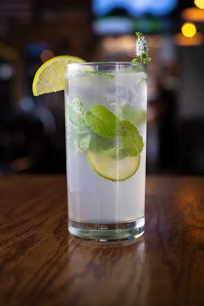 Tall cocktail with mint, lime, flower, and condensation on wood.
