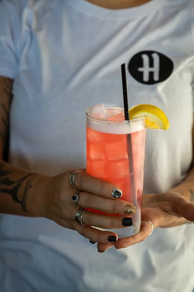 Tattooed hands with painted nails hold a pink cocktail with lemon.