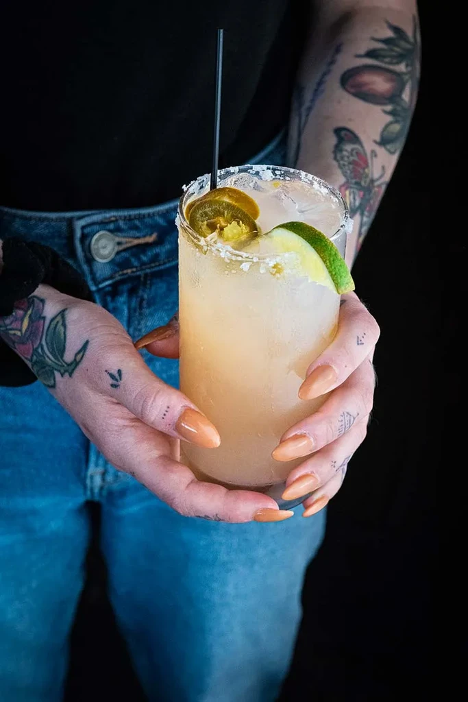 Tattooed hands holding a jalapeño cocktail with lime and salted rim.