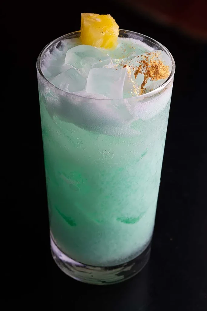 Frothy light green cocktail with ice, pineapple garnish, and golden glitter.