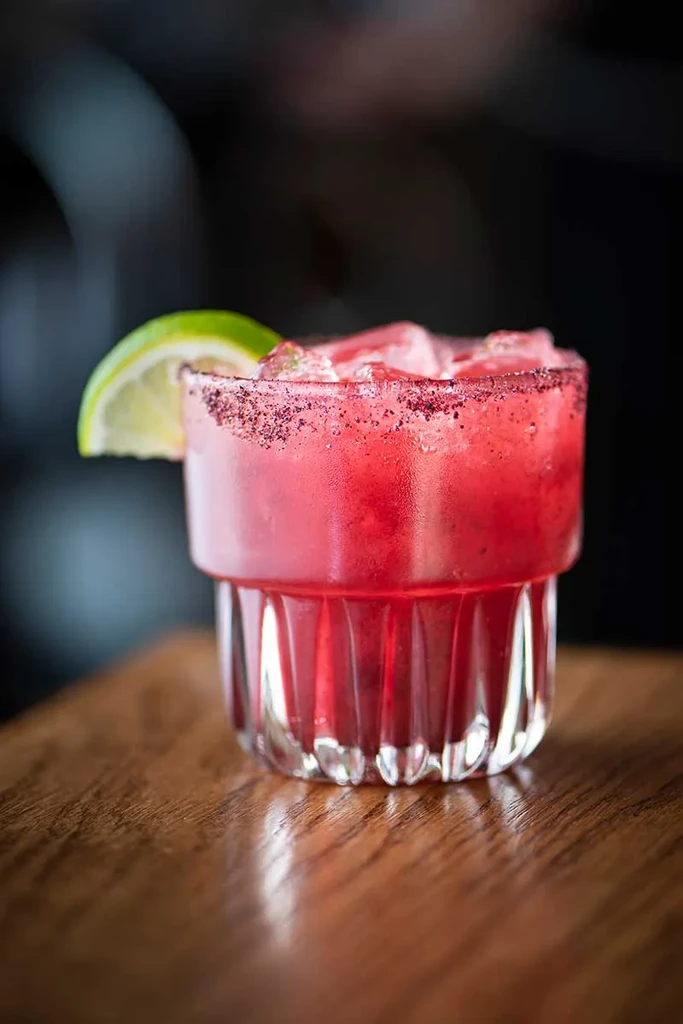 Deep red cocktail with ice, dark rim, lime, on wood.