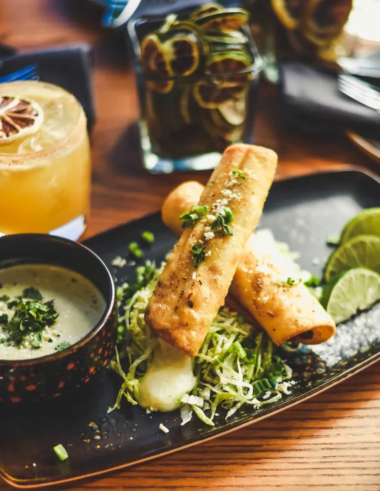 Two garnished taquitos, green dip, and cocktails on a wooden table.