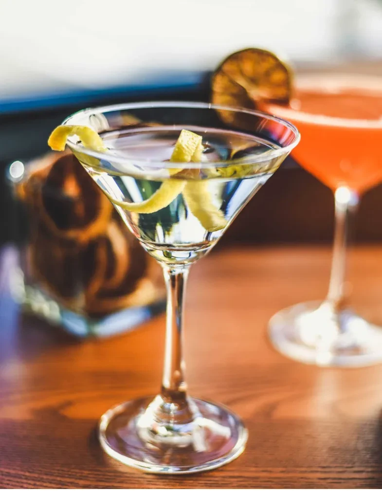 Clear martini with lemon twist, blurred red cocktail on wood.