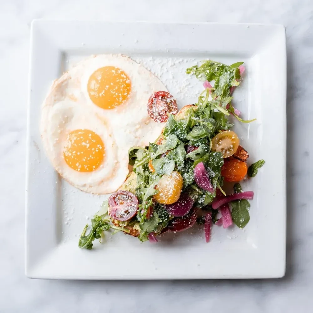 Top-down: Fried eggs, arugula, tomatoes, pickled onions on a white plate.