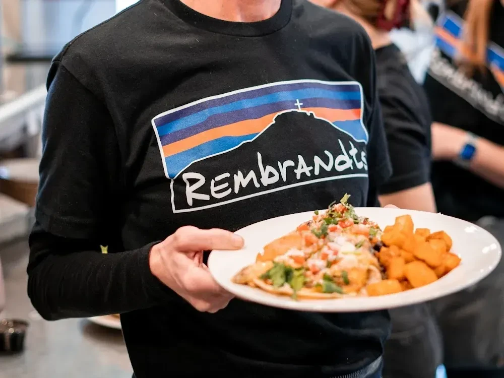 Person holding a plate of tacos and fries, wearing a "Rembrandt's" shirt.