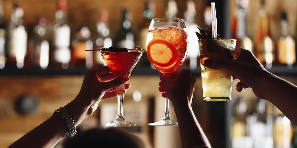 Three hands toast with varied cocktails at a blurred bar.