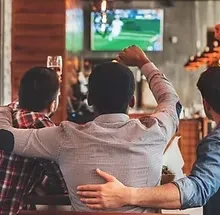 Three men at a bar watch sports; one cheers with raised fist.