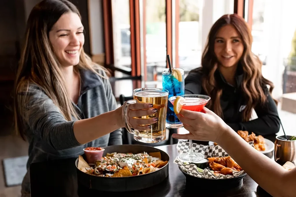 Three people toast with beer and cocktails over nachos and wings.