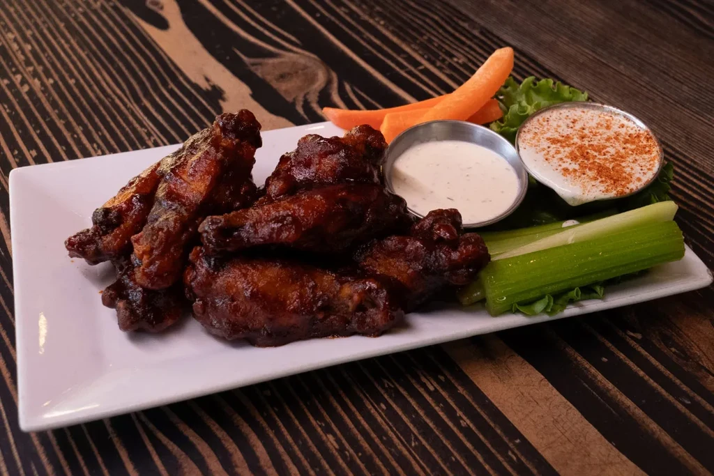 Plate of glistening barbecued chicken wings with sauces, celery, and carrots.
