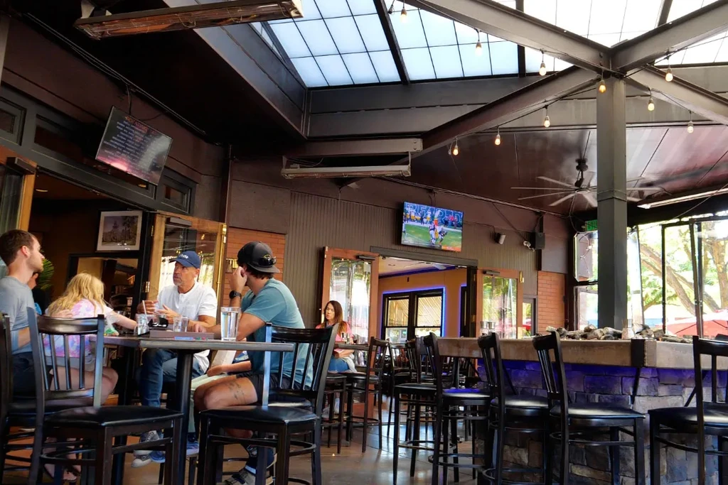 Restaurant patio under skylight, exposed beams, with TVs and patrons.