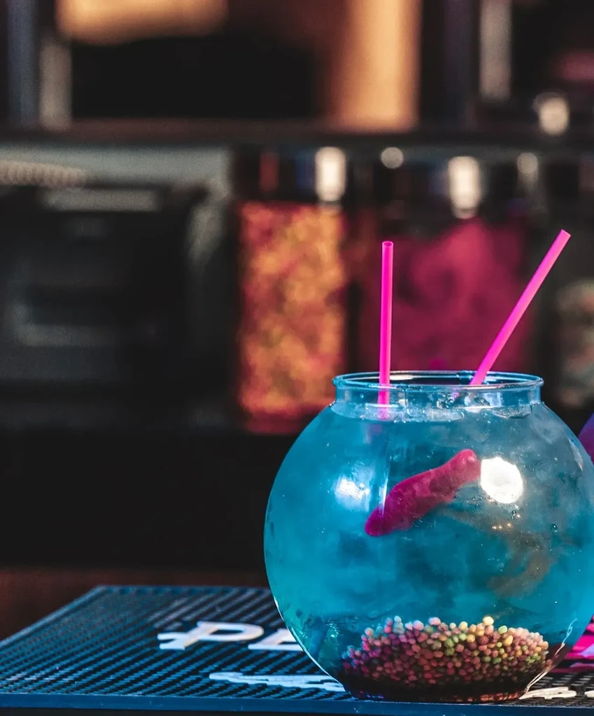 Blue drink in fishbowl with pink straws, red gummy, and colorful candies.