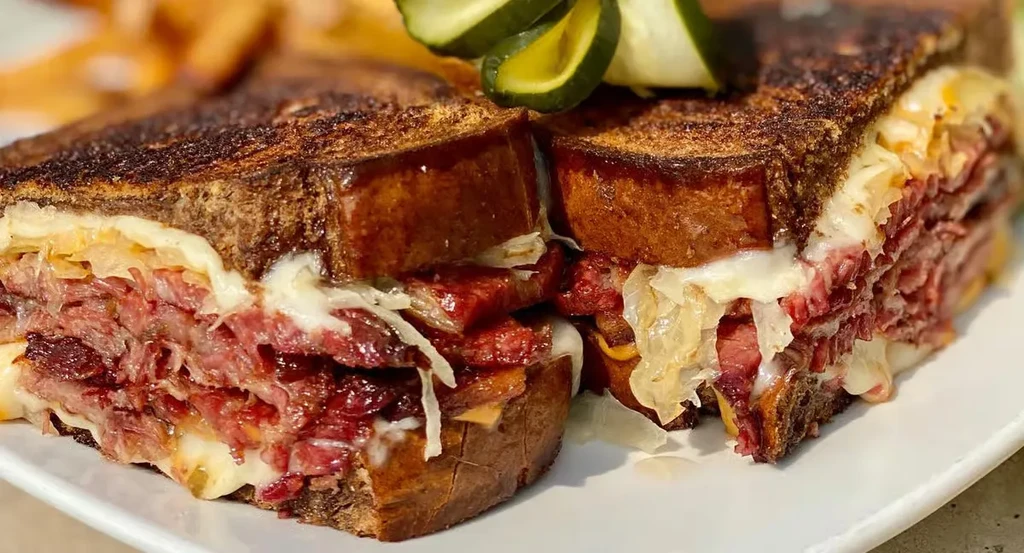 Large Reuben sandwich cut in half with pickles and french fries.