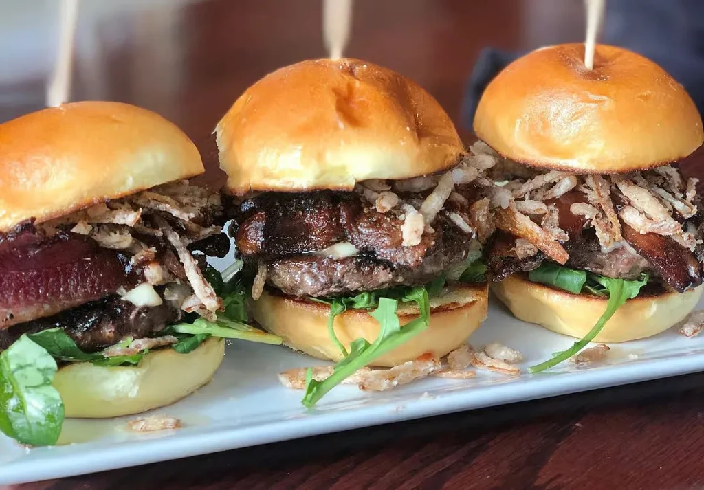 Three brioche sliders with patties, arugula, crispy onions, and bacon.