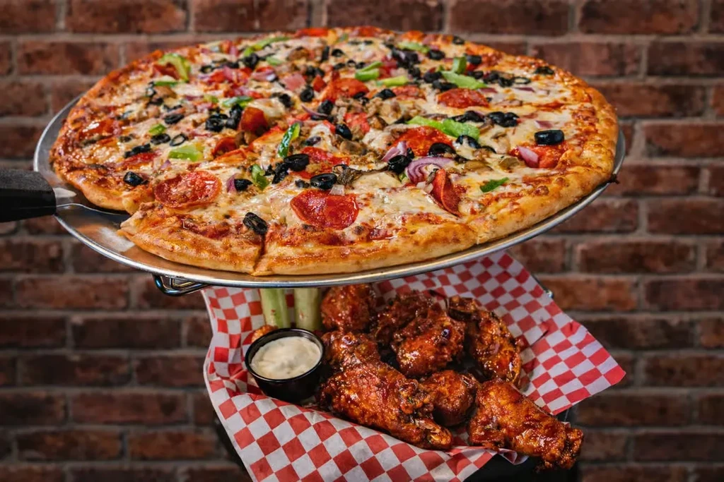 Pizza on a peel above chicken wings, sauce, and celery.