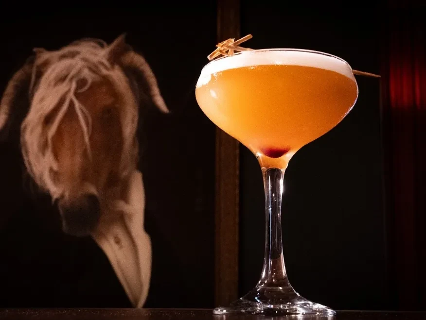 Bright orange cocktail with frothy head, wooden garnish, skull behind.