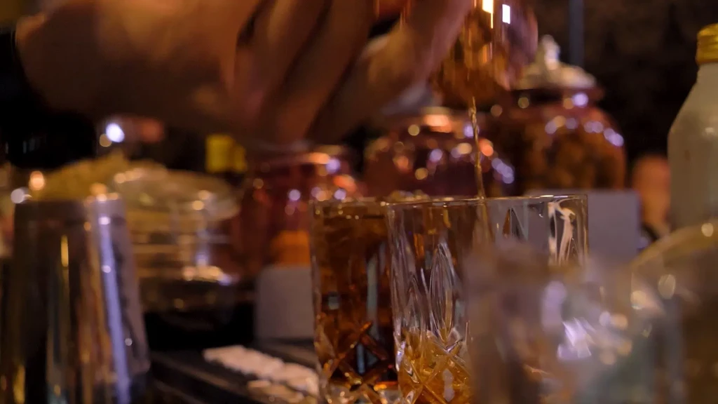 Bartender pours golden spirit from jigger into two ornate tumblers.