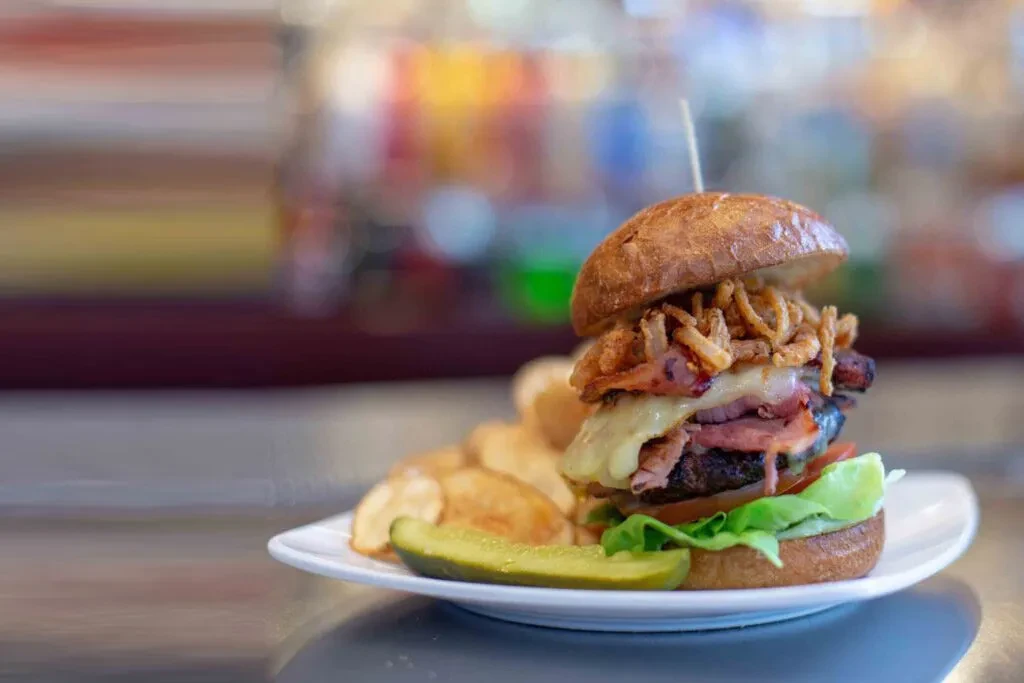 Tall gourmet burger with melted cheese, crispy onions, chips, and a pickle.