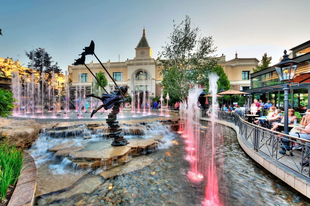 Illuminated fountain with bronze girl sculpture, restaurant patio at dusk.