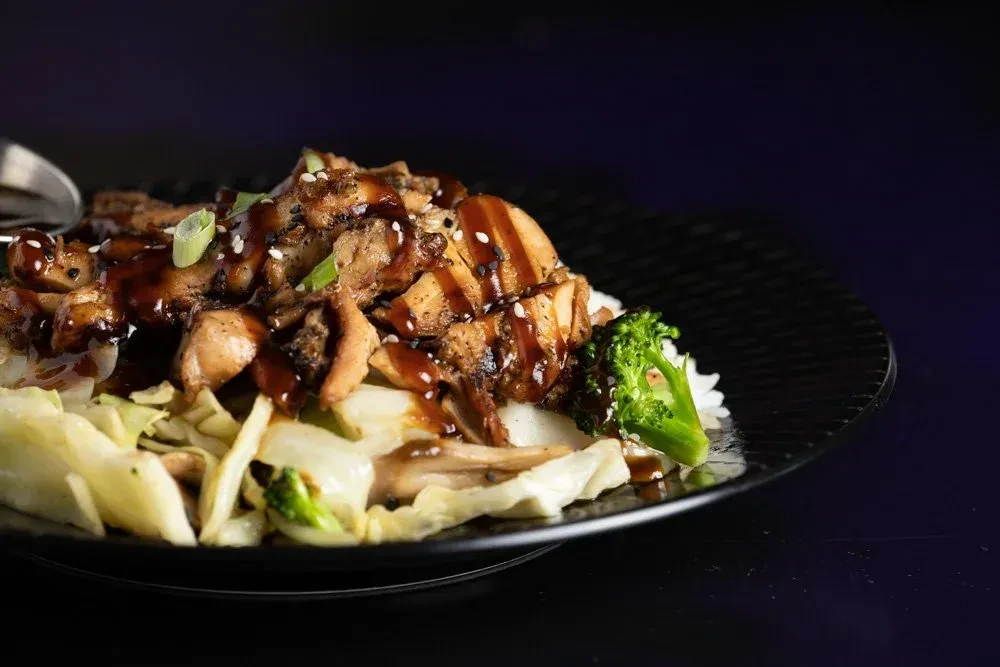 Close-up of sauced chicken, rice, cabbage, and broccoli on a dark plate.