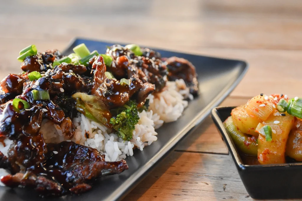 Dark-glazed meat, rice, broccoli, and kimchi-like side on a black plate.