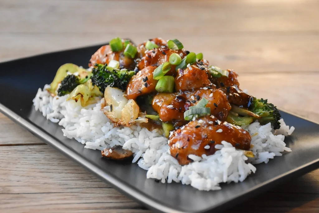Glazed chicken, rice, broccoli, sesame seeds on a black plate.