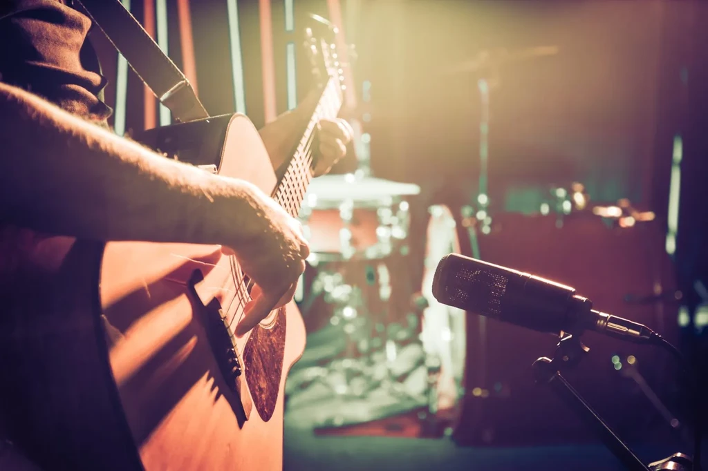 Musician's hands strumming an acoustic guitar under warm stage lights.