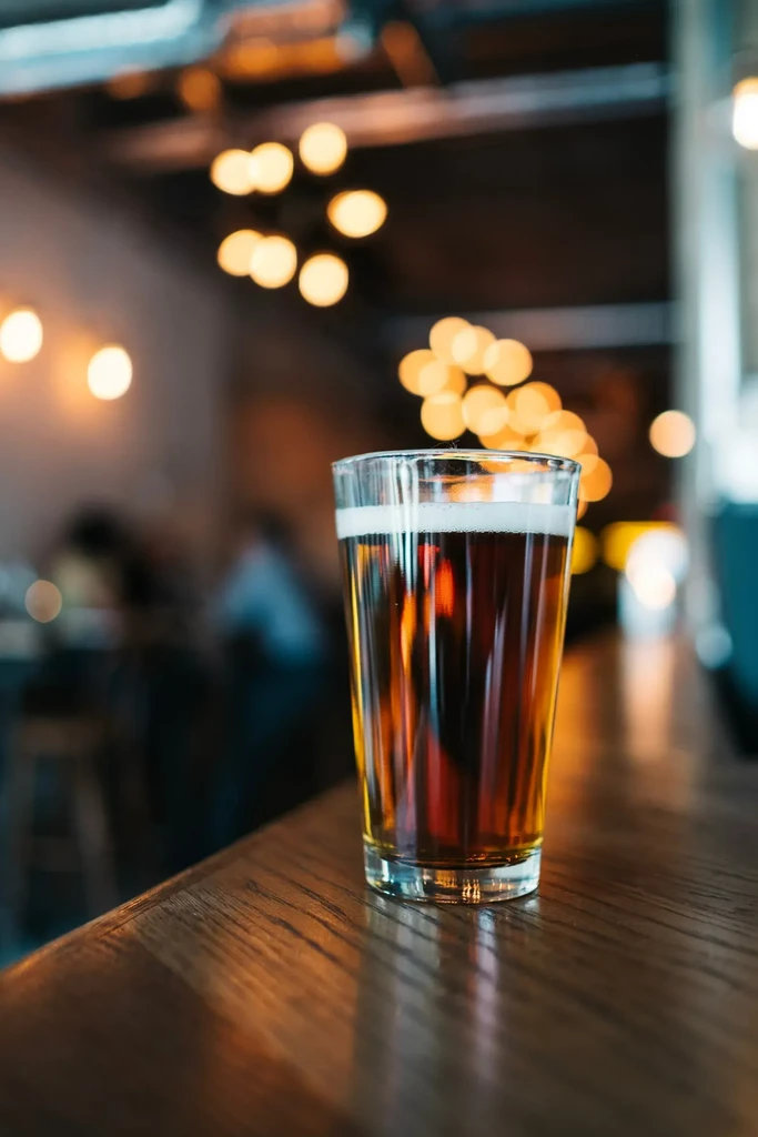 Glass of dark amber beer on a wooden bar with blurred warm lights.