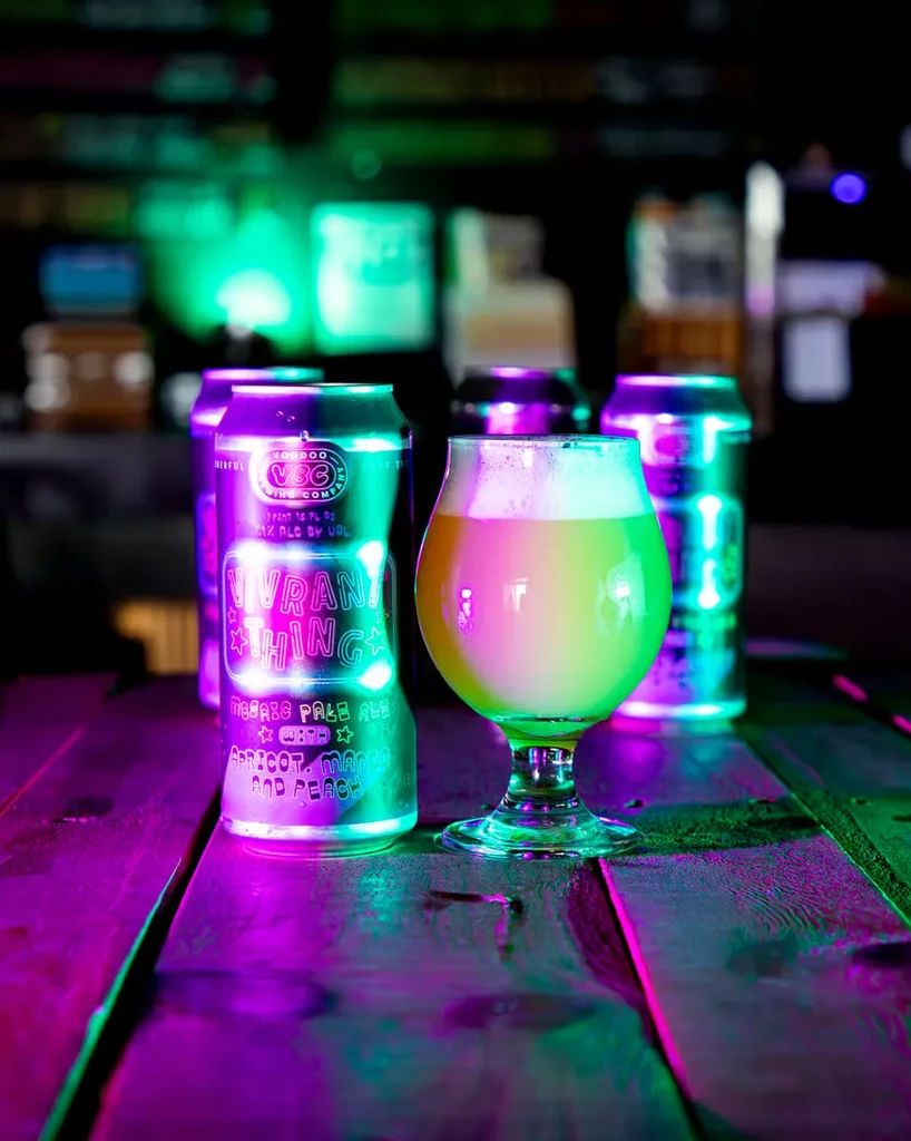 Close-up of hazy beer in glass, with neon-lit Voodoo Brew cans.