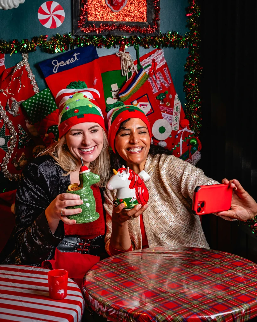 Smiling women in Christmas hats hold novelty drinks, one taking a selfie.