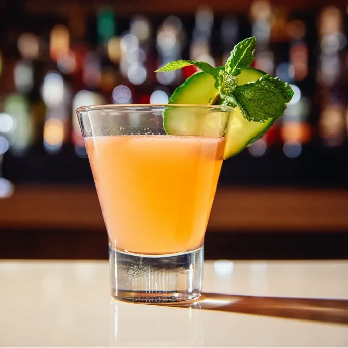 Reddish-orange cocktail with cucumber and mint on a bar counter.