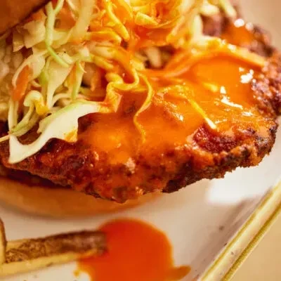 Close-up fried chicken sandwich with orange hot sauce and coleslaw on plate.