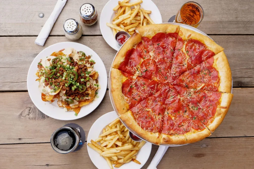 Overhead view: pepperoni pizza, nachos, fries, and drinks on a rustic table.