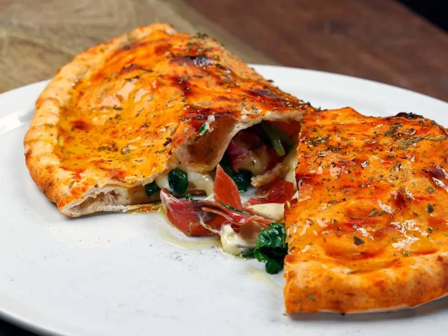 Golden-brown calzone, cut to reveal melted cheese, spinach, and cured meat.