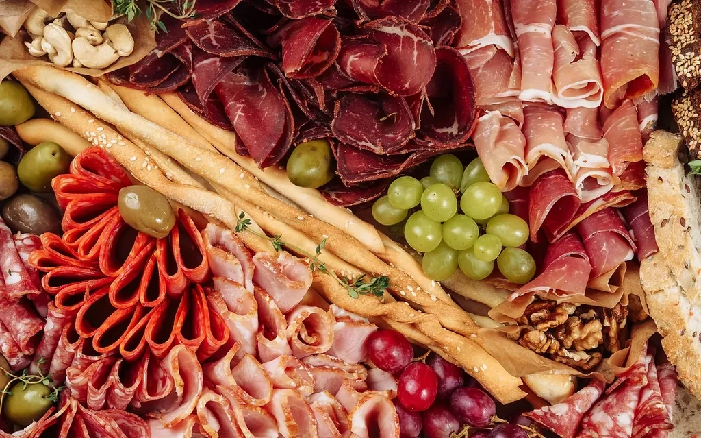 Top-down charcuterie and cheese board with meats, fruits, nuts, bread.