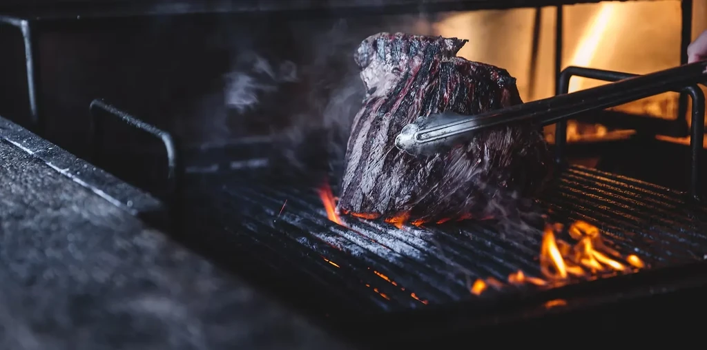 Steak grilled over open flame, held by tongs, actively steaming.