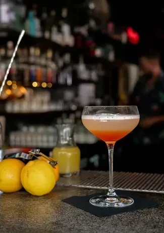 Layered cocktail in coupe glass on bar with citrus and blurry bartender.