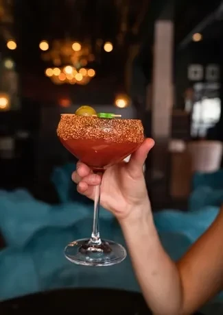 Red cocktail with seasoned rim and olive, held in hand.