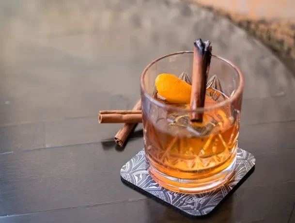 Amber cocktail in faceted glass with charred cinnamon, orange peel.
