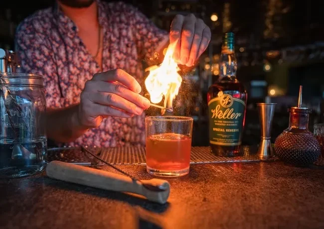 Bartender's hands ignite a flaming amber cocktail at a dimly lit bar.