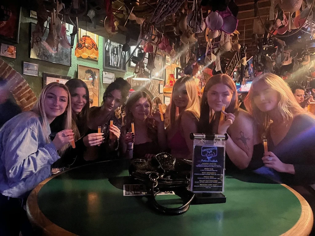 Six smiling women drink test-tube cocktails under a bra-adorned ceiling.