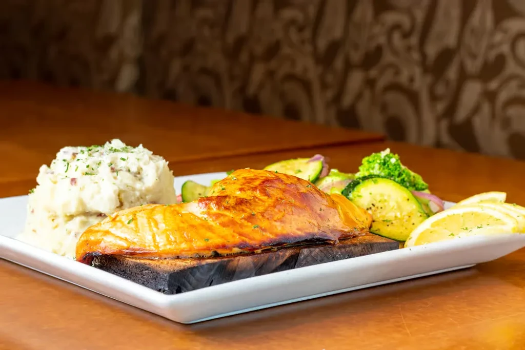 Cedar plank salmon, mashed potatoes, sautéed vegetables, and lemon wedges.